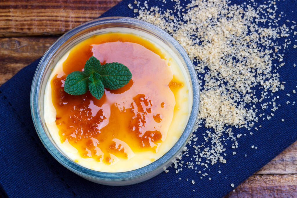 This vibrant image captures the essence of a classic French crème brûlée, featuring the iconic rich caramel layer that has been expertly caramelized to a shiny, amber finish. A fresh sprig of mint rests atop, adding a pop of color and hint of freshness. Beside the ramekin, a scattering of coarse sugar crystals on a deep blue napkin suggests the sugar's transformation into the dessert's alluring, crackled top. The rustic wooden background adds warmth to the composition, invoking the cozy ambiance of a French bistro.