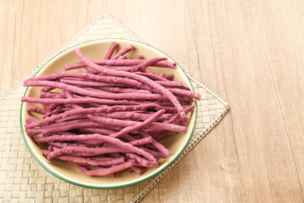 A bowl of thin-cut purple sweet potato sticks on a beige woven mat placed on a light wood surface, highlighting the vibrant hue of the healthy tuber.