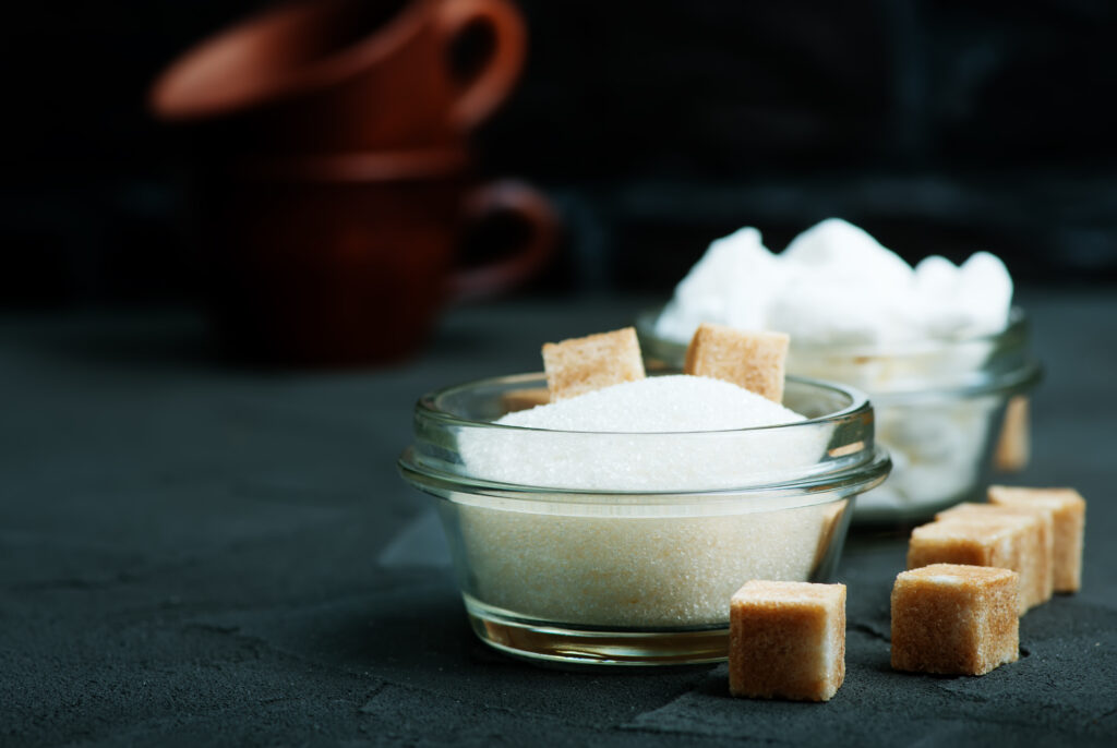 This image artfully displays a clear glass bowl filled with granulated white sugar, positioned on a dark, textured surface that accentuates the sugar's purity and crystalline texture. Adjacent to the bowl, perfectly shaped brown sugar cubes offer a contrast in color and texture, suggesting a variety of sweetening choices. In the background, soft focus hints at a warm kitchen setting, possibly with terracotta pots, creating a cozy atmosphere for baking and culinary creations.