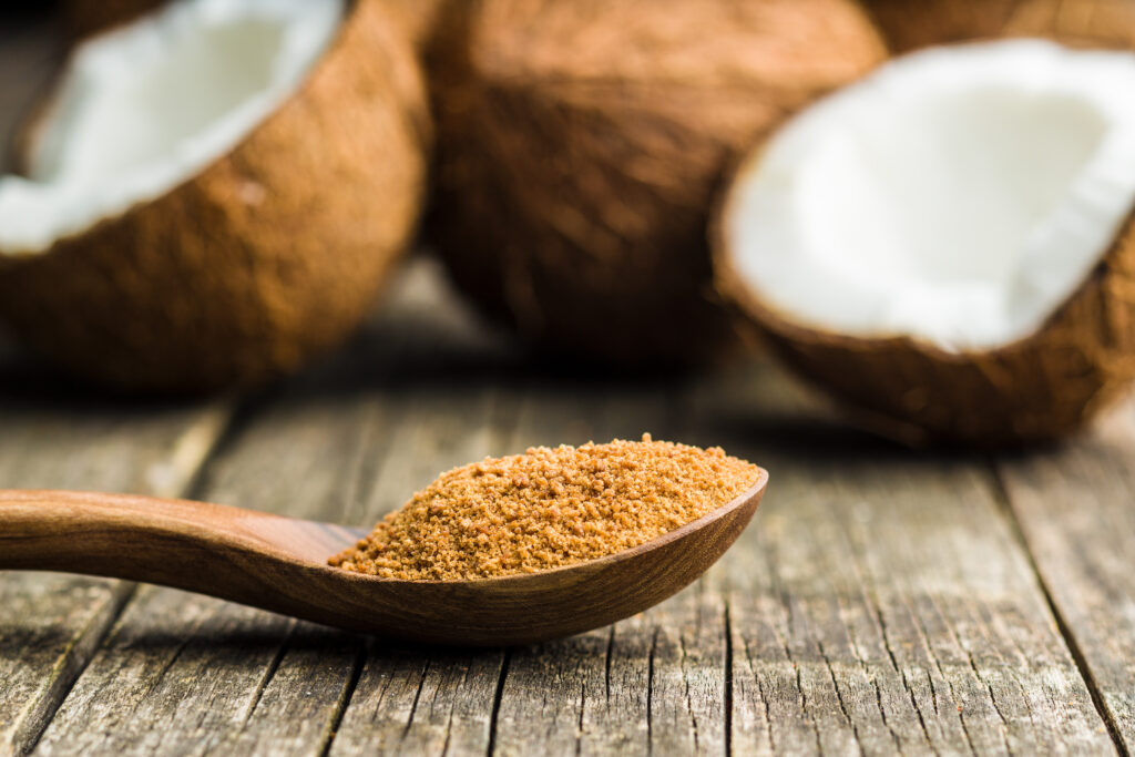 This inviting image captures the natural essence of coconut sugar resting in a wooden spoon, its caramel hues rich and warm against the rustic backdrop. In the background, halved coconut shells add a tropical touch, evoking the sugar’s exotic origins. The texture of the sugar is finely granular, suggesting its unrefined nature and the robust flavor it could lend to sweet and savory dishes alike. The old wood surface it lies on tells a story of traditional cooking, bridging the gap between old-world methods and modern culinary adventures.
