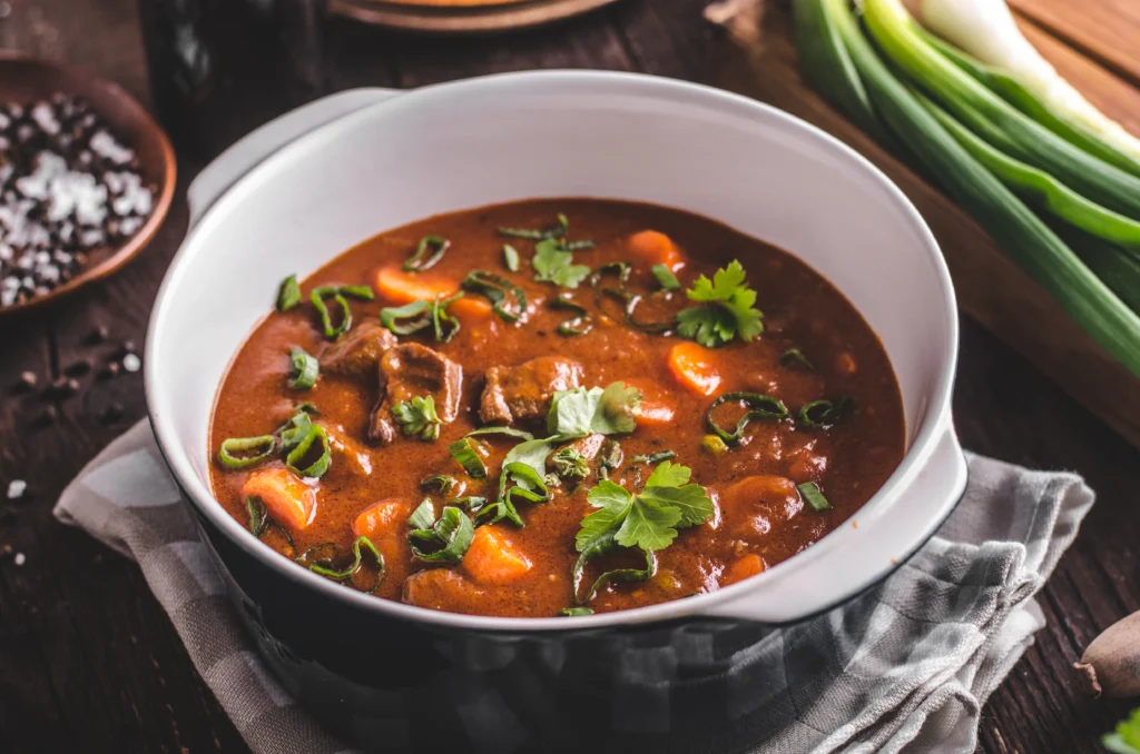This image features a hearty beef stew, brimming with tender chunks of beef, vibrant carrots, and potatoes, all simmered to perfection in a rich, savory broth. The stew is garnished with fresh herbs, adding a burst of color and flavor that enhances the visual appeal and taste of this classic comfort dish.
