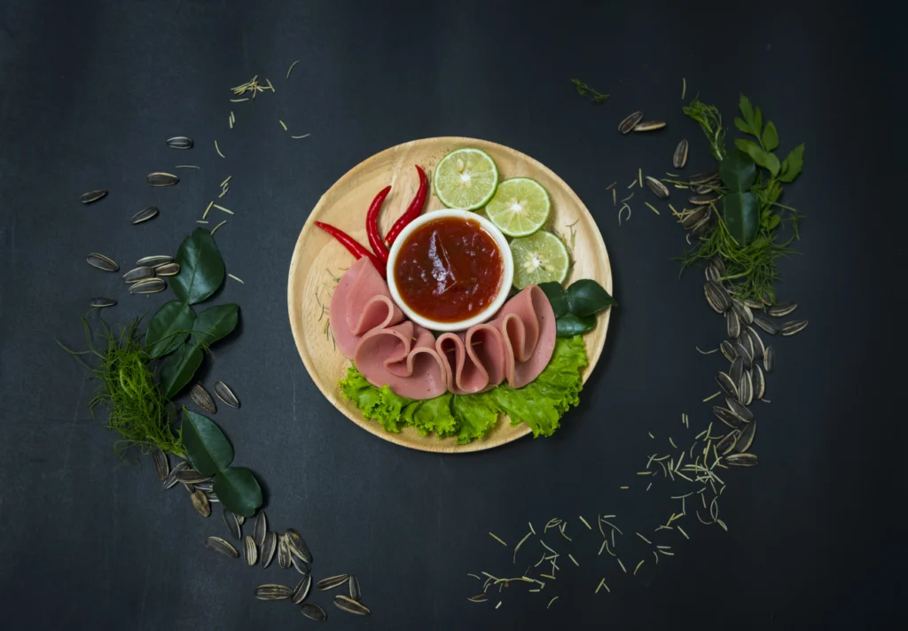This image captures a stunning culinary presentation of bologna, thoughtfully arranged on a wooden plate to enhance its visual appeal. The bologna slices are delicately folded, surrounded by fresh green lettuce leaves, and accompanied by slices of lime, red chili peppers for a touch of spice, and a small bowl of rich red sauce for dipping. Scattered around the plate are various fresh herbs and sunflower seeds, adding texture and depth to the arrangement. Set against a sleek, dark background, the vibrant colors and meticulous plating transform the simple bologna into a gourmet dish, perfect for those looking to appreciate a creative and flavorful approach to traditional ingredients.