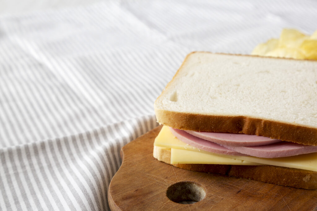 A homemade bologna and cheese sandwich on white bread, neatly placed on a wooden cutting board with a striped tablecloth background.