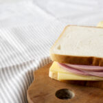 A homemade bologna and cheese sandwich on white bread, neatly placed on a wooden cutting board with a striped tablecloth background.