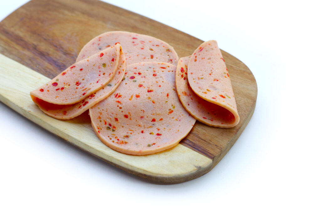 The image shows several slices of spicy bologna arranged neatly on a wooden cutting board. The bologna is speckled with visible bits of red and green spices, indicating a seasoned variety that likely offers a peppery flavor. The backdrop is a stark white, highlighting the vivid colors and textures of the bologna, making it visually appealing and suggesting a readiness to be used in cooking or served as part of a meal. The wooden board adds a rustic touch that complements the simple yet flavorful appearance of the sliced bologna.